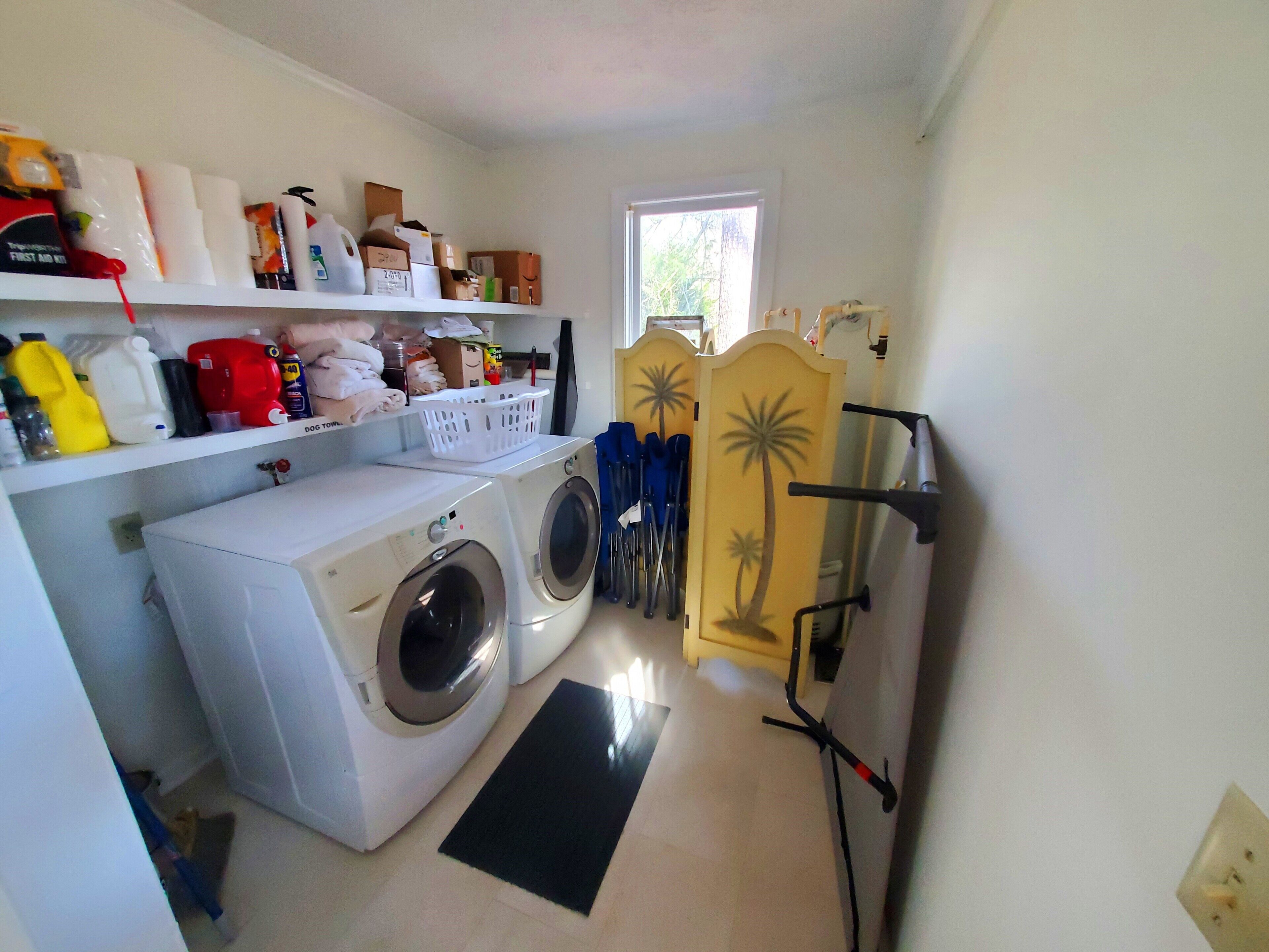 A laundry room with a front-loading washer and dryer, shelves with cleaning supplies, a window, and a door with a palm tree design.