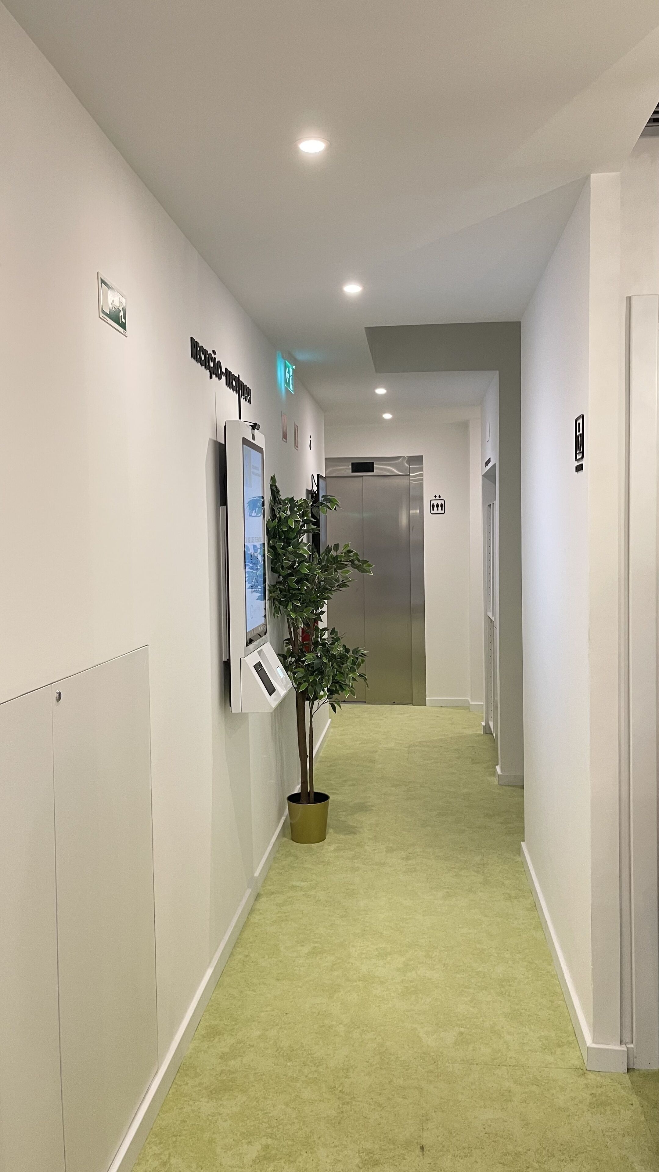 A hallway with a reception desk, a digital information kiosk, a potted plant, and an elevator.