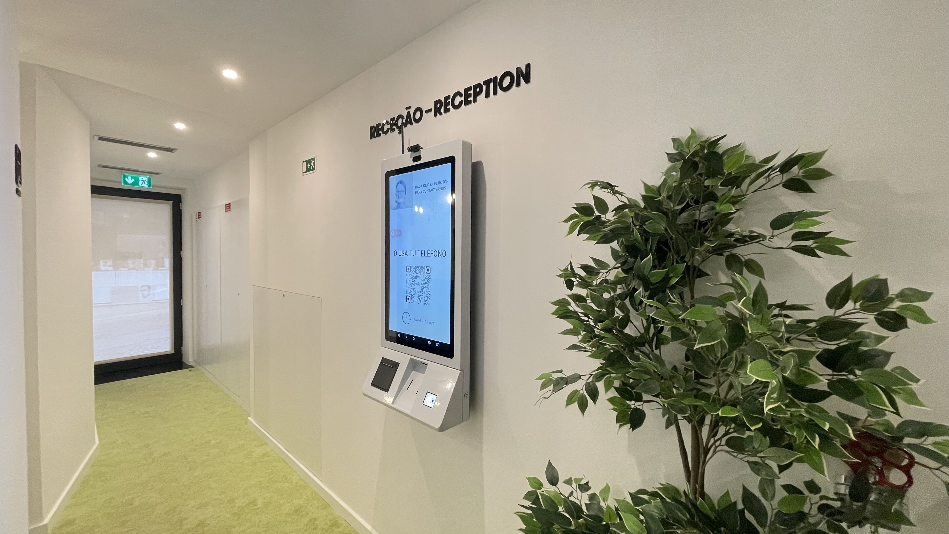 A reception area with a digital kiosk, a green carpeted floor, and a potted plant.
