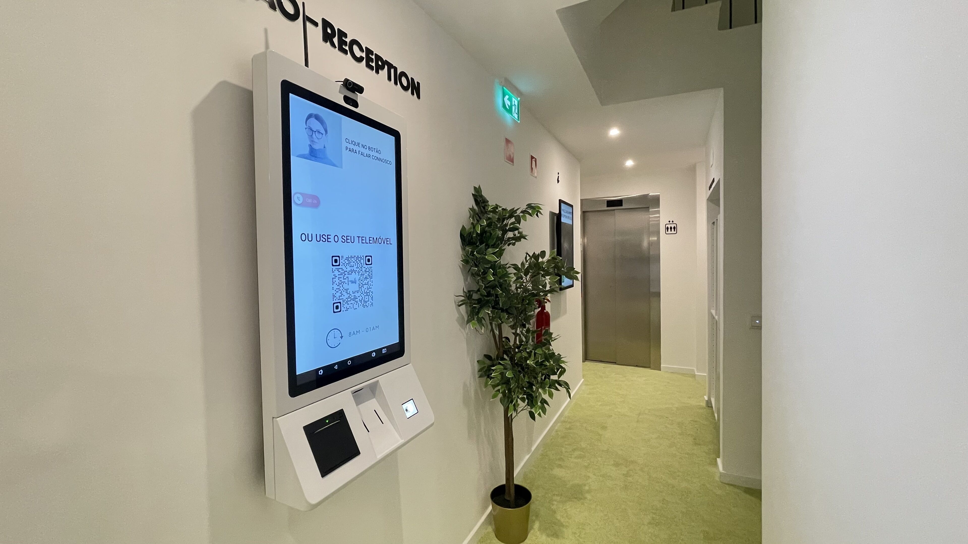 Uma área de recepção com um quiosque digital, uma placa de saída verde, um elevador e um vaso de plantas.