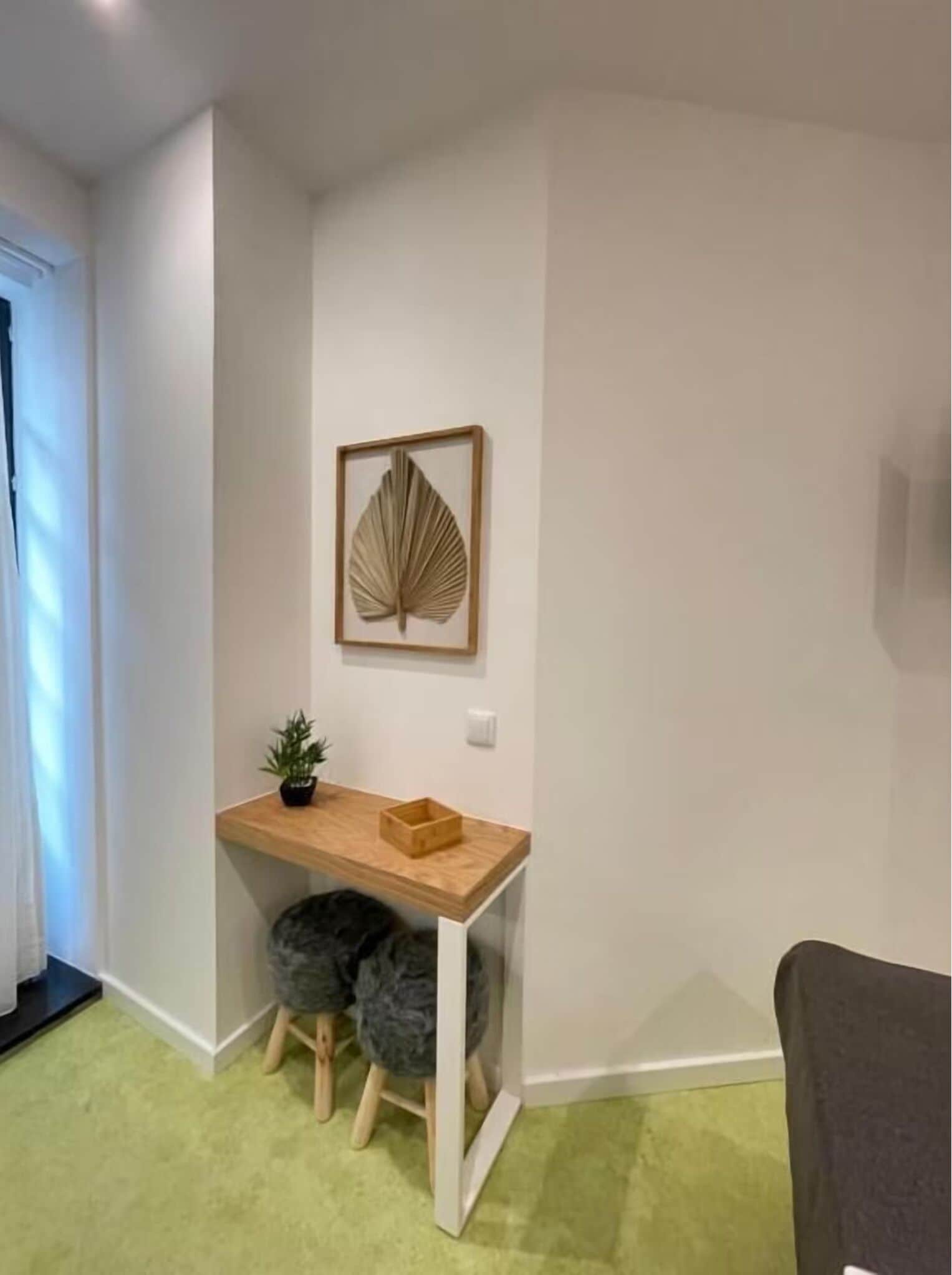Um quarto compacto com uma escrivaninha de madeira, um banquinho pequeno, uma obra de arte emoldurada e um vaso com planta.