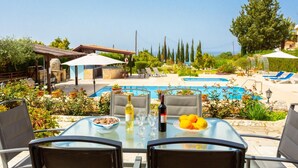 Un área de comedor al aire libre con piscina, sillas y una mesa dispuesta con copas de vino y fruta.