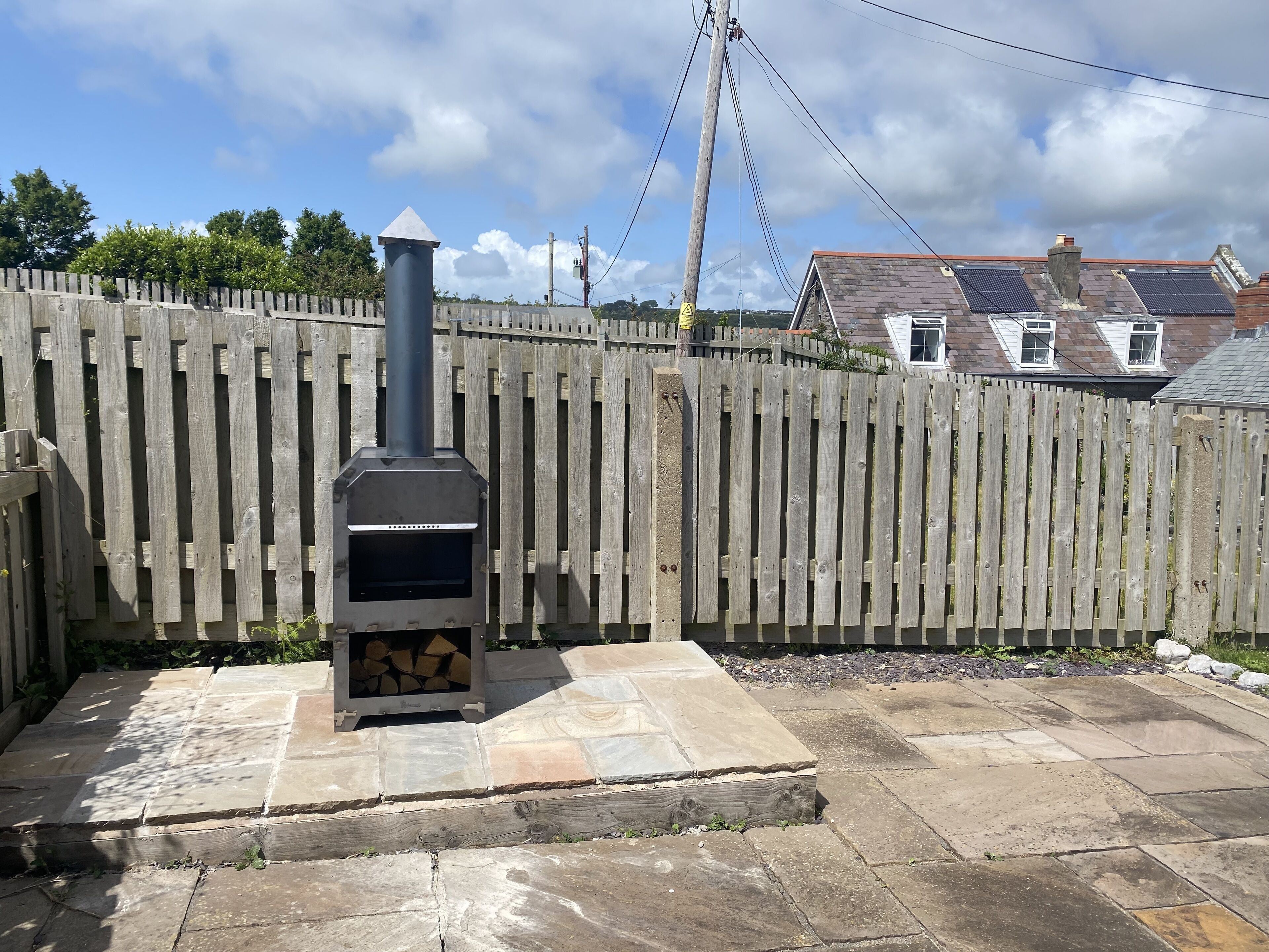 A stone patio with a wood-burning stove, a wooden fence, and houses in the background.