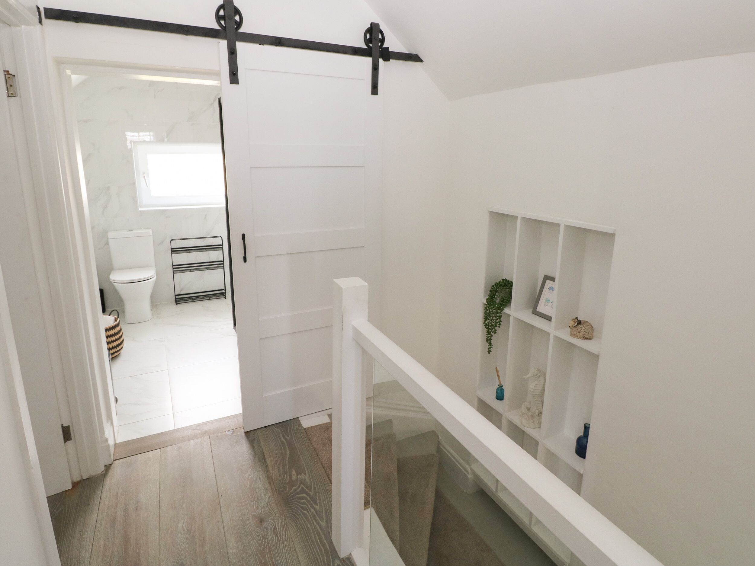 Ein Korridor mit einer weißen Tür zu einem Badezimmer, einem Holzboden und einem weißen Geländer mit Regalen.