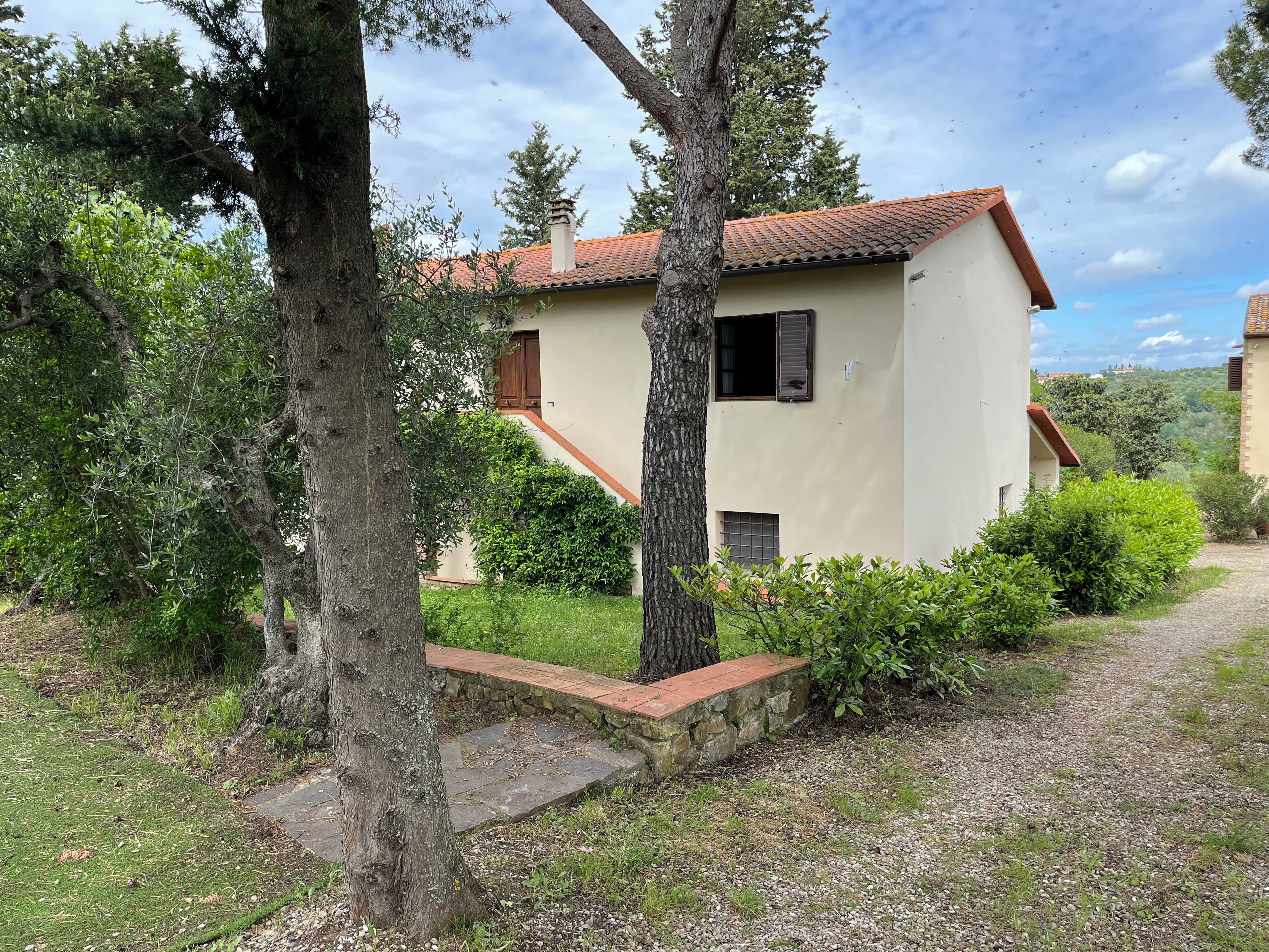 Une maison à deux étages, avec un toit recouvert de tuiles rouges, entourée de verdure et d’arbres.
