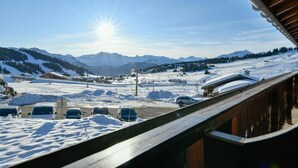 Et sneklædt bjerglandskab med klar himmel, en træbygning med balkon, og flere parkerede biler.