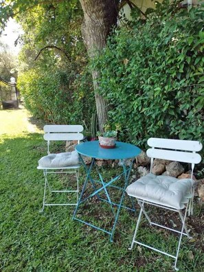 Un petit espace extérieur aménagé avec deux chaises en métal blanc et une table turquoise, le tout entouré d’une végétation luxuriante et d’un arbre.