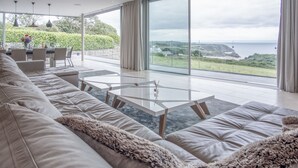 A modern living room with a large sofa, a glass coffee table, and a view of the sea. - Bryn House - 5 Bed Home - Penmaen (Swansea)