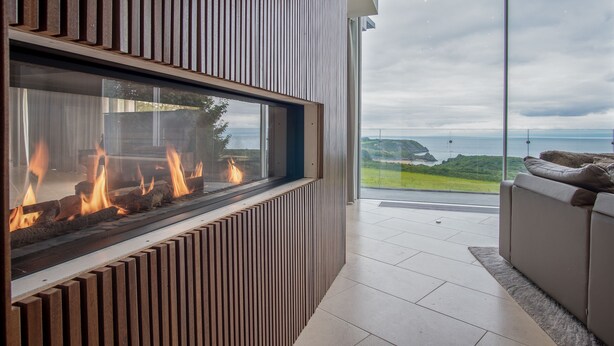 A modern living room with a fireplace, a sofa, and a view of the ocean. - Bryn House - 5 Bed Home - Penmaen (Swansea)
