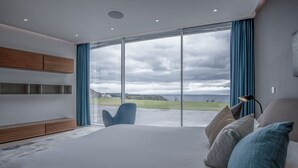 A modern bedroom with a large bed, a blue chair, and a view of the sea. - Bryn House - 5 Bed Home - Penmaen (Swansea)