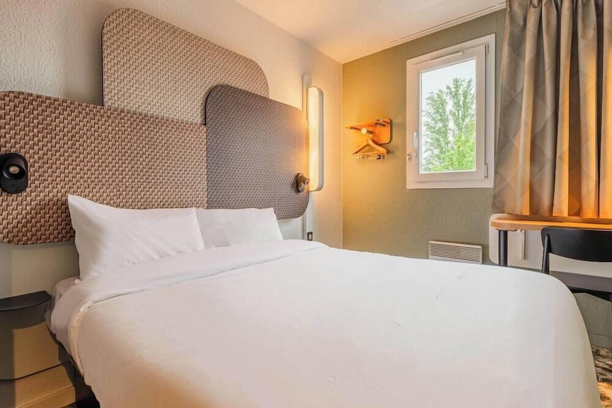 Et hotelværelse med en stor seng, en vægmonteret lampe, et vindue med gardiner og et lille bord med en stol.