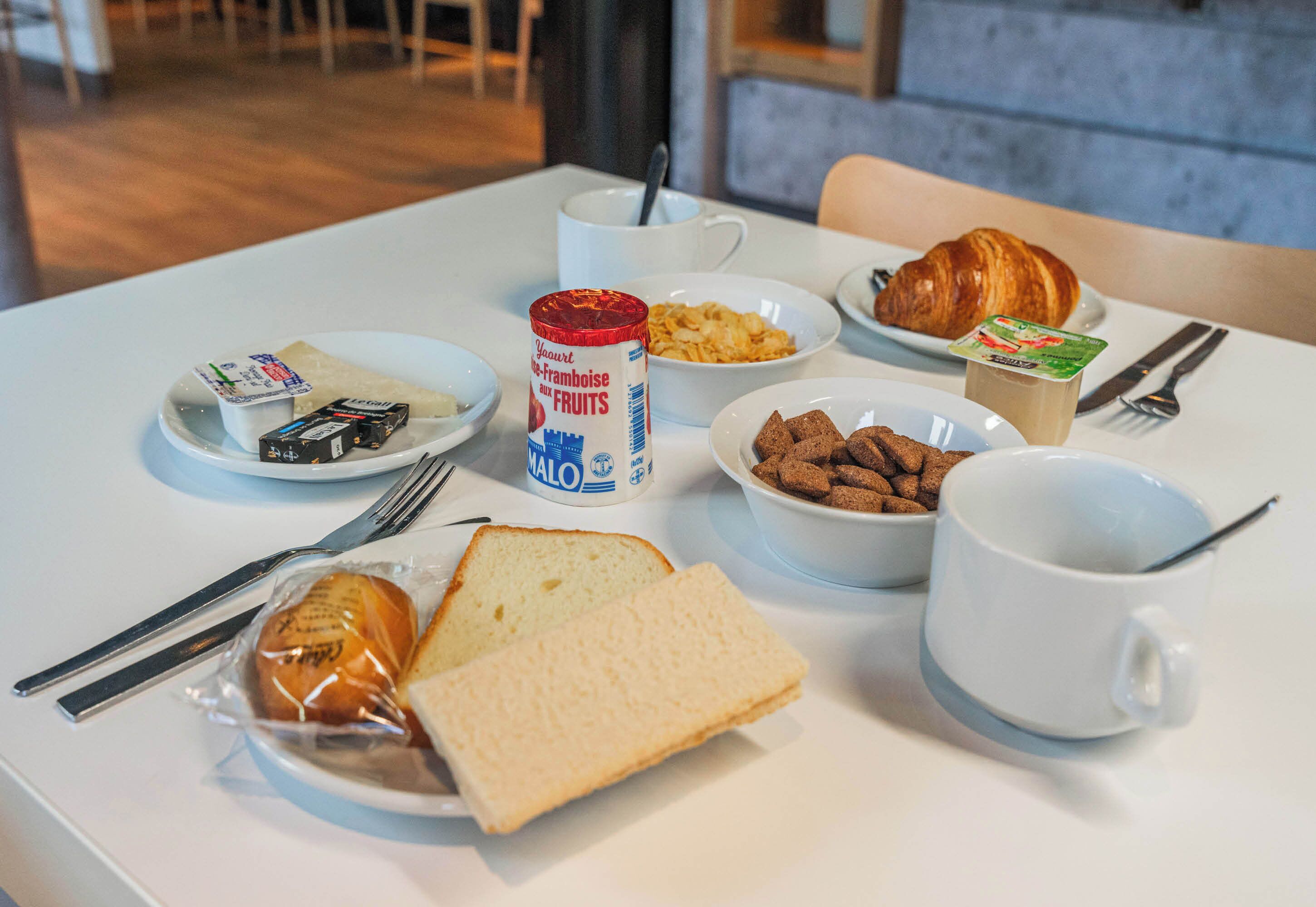 Una mesa de desayuno con croissants, pan, cereal, yogur y otro croissant sobre un mantel blanco.