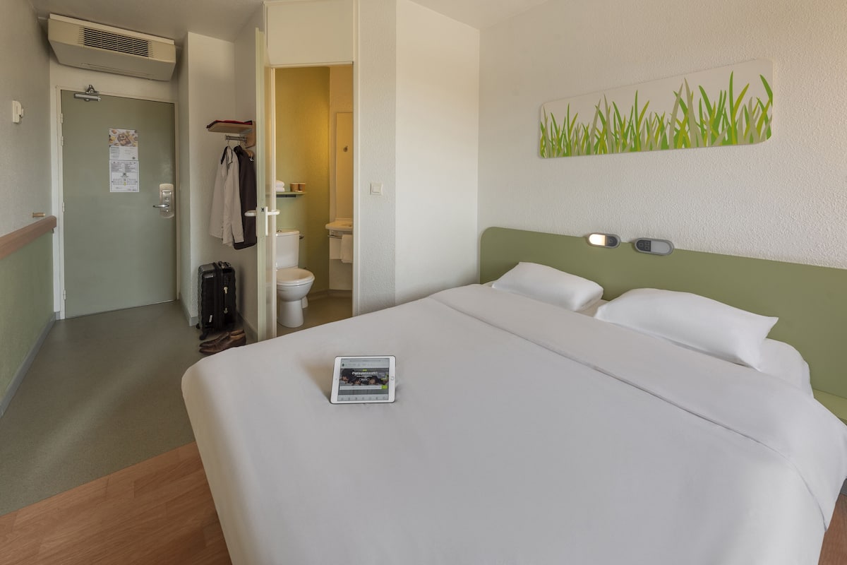 Une chambre d’hôtel comprenant un lit, une table de chevet avec une tablette, une lampe fixée au mur et une porte donnant accès à la salle de bain.