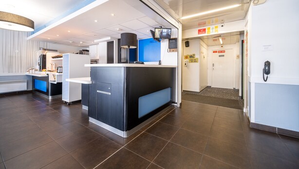 A modern reception area with a counter, a blue wall, and a door marked "Exit". - B&B HOTEL Calais Terminal Cité de l'Europe 2 étoiles (Coquelles)