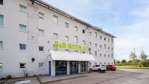 A B&B hotel with a white facade, multiple windows, and a parking area with two cars. - B&B HOTEL Calais Terminal Cité de l'Europe 2 étoiles (Coquelles)