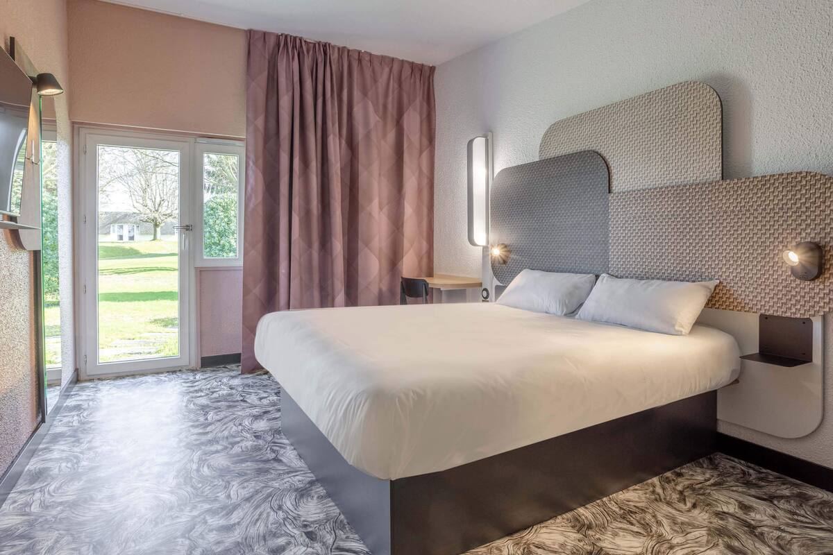 Une chambre à coucher moderne avec un grand lit, une porte-fenêtre coulissante donnant sur l’extérieur et une vue sur une pelouse verdoyante.