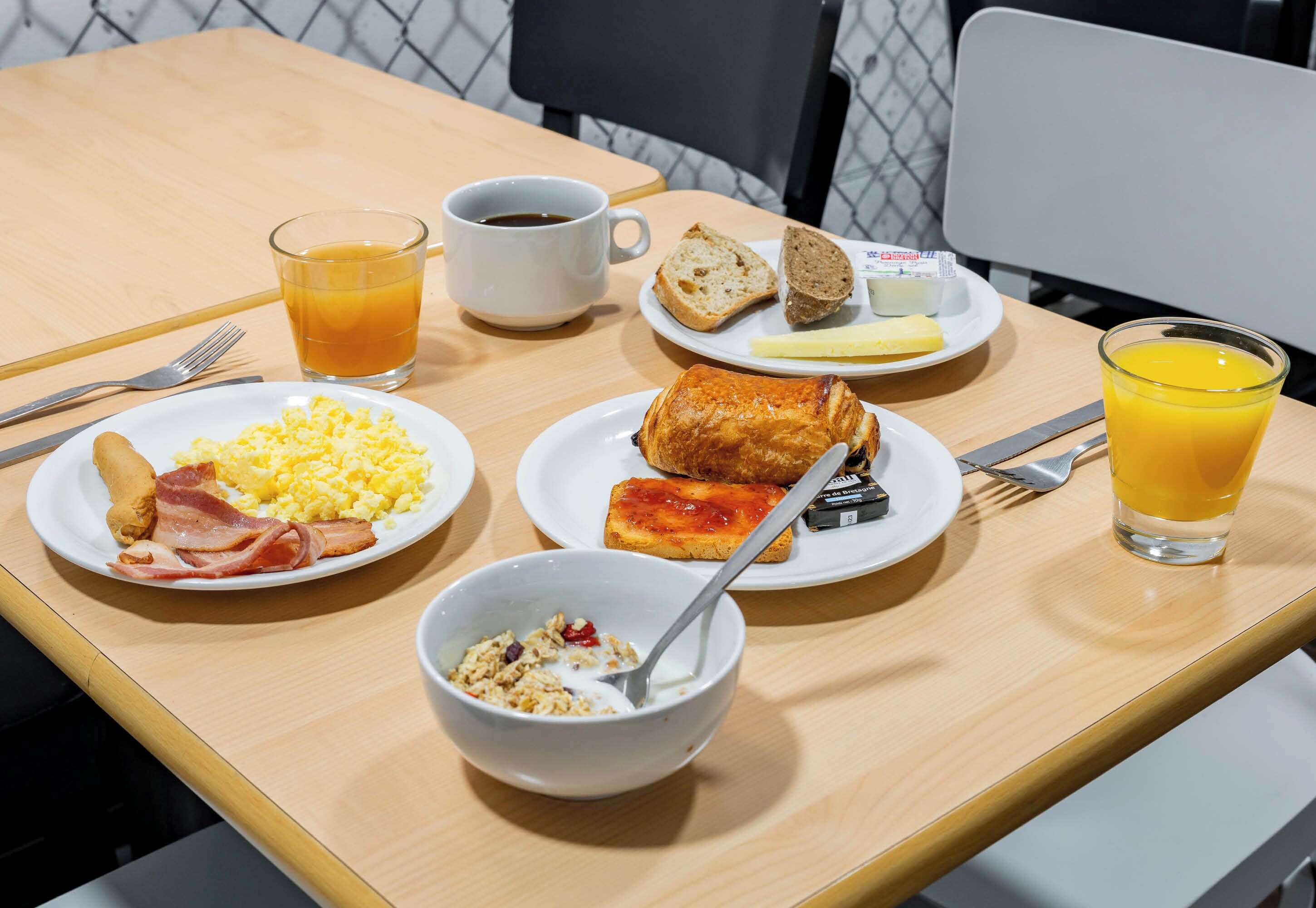 A breakfast table with plates of eggs, bacon, toast, and a bowl of cereal. There are also cups of coffee and orange juice.