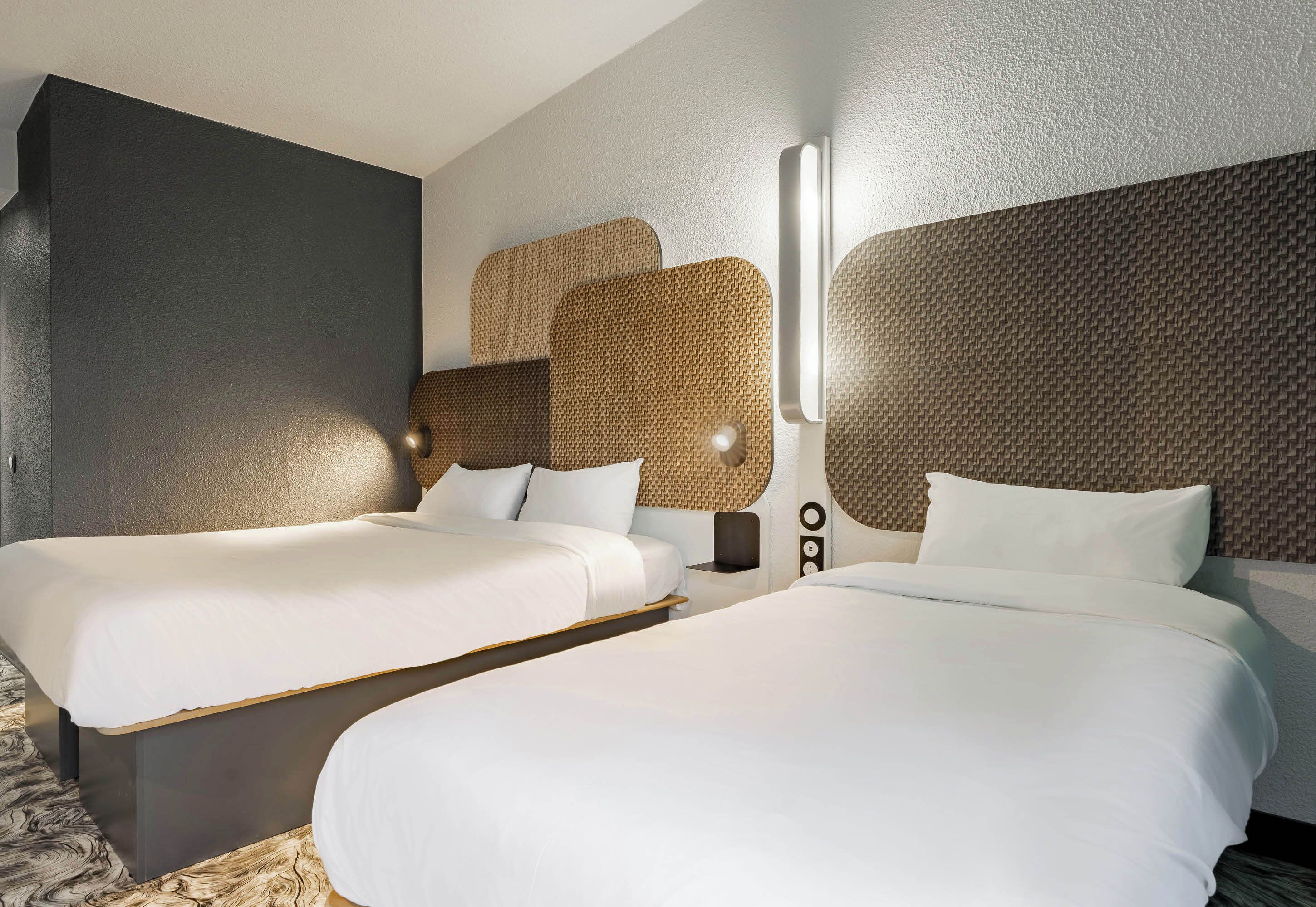 Quarto de hotel com duas camas, luminária grande fixada na parede e design texturizado na parede.