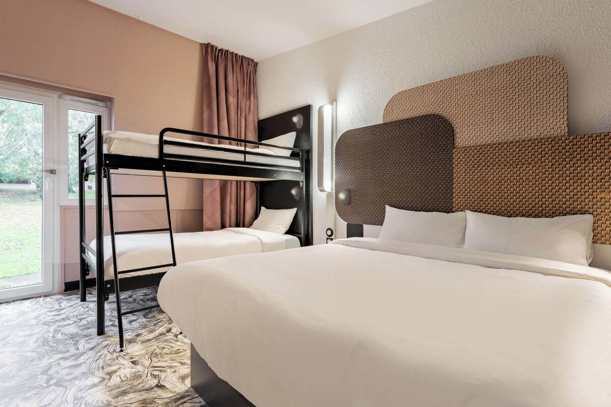 Une chambre d’hôtel avec un lit superposé, un lit séparé, une grande fenêtre avec des rideaux et une tête de lit en osier.