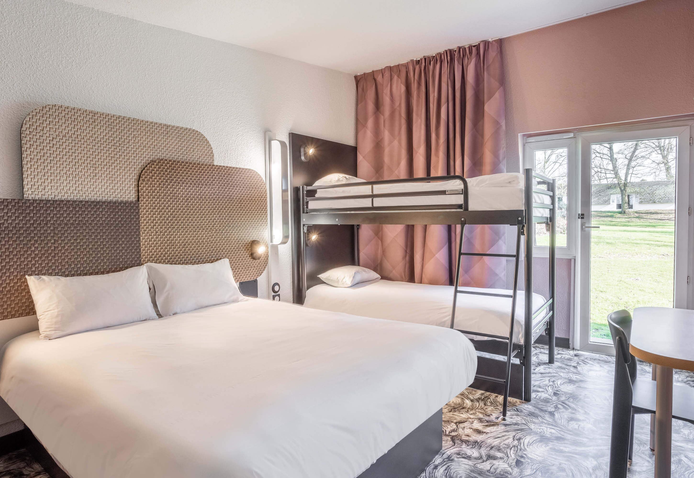 A hotel room with a bunk bed, a single bed, a desk, and a large window with curtains.