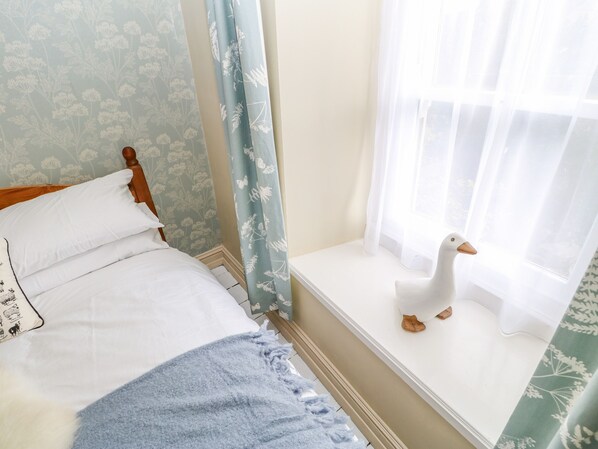 Une chambre à coucher avec un lit, un coin fenêtre agrémenté d’une décoration en forme de canard blanc, un papier peint à motifs floraux et un rideau bleu.