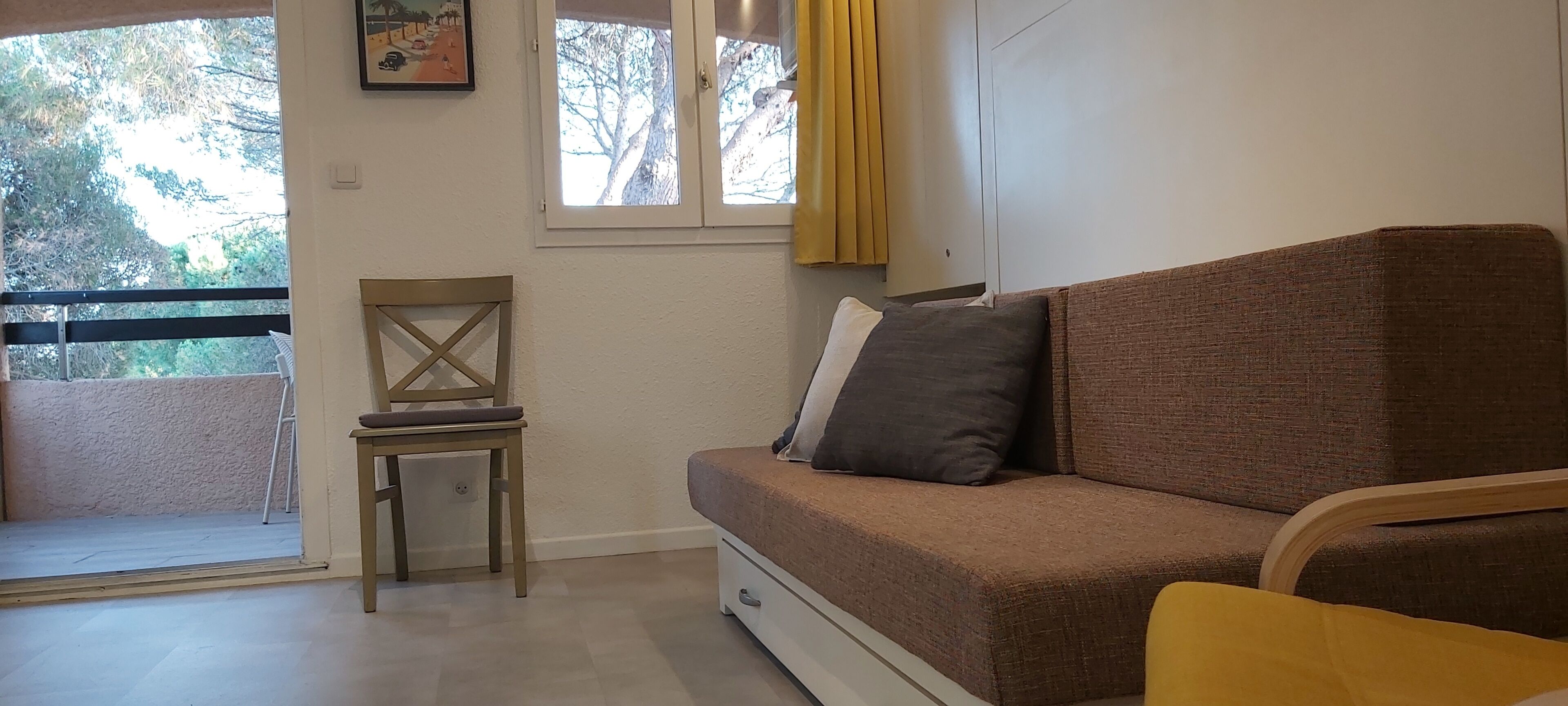 Una habitación con sofá marrón, una silla de madera, una mesita, una ventana con cortinas y una puerta que da a un espacio al aire libre.