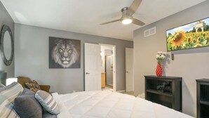 Une chambre avec un grand lit, un ventilateur de plafond, un tableau représentant un lion, une télévision et une porte donnant sur une salle de bain.