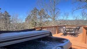Un bain à remous sur une terrasse en bois, avec vue sur les arbres, et un espace patio aménagé avec des chaises et une table.