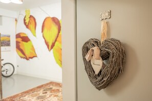 A room with a patterned rug, a wall-mounted heart-shaped decoration with a tag, and a wall with a painting of birds.
