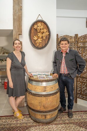 Un hombre y una mujer de pie junto a un barril de madera, con una pared rústica y detalles decorativos al fondo.