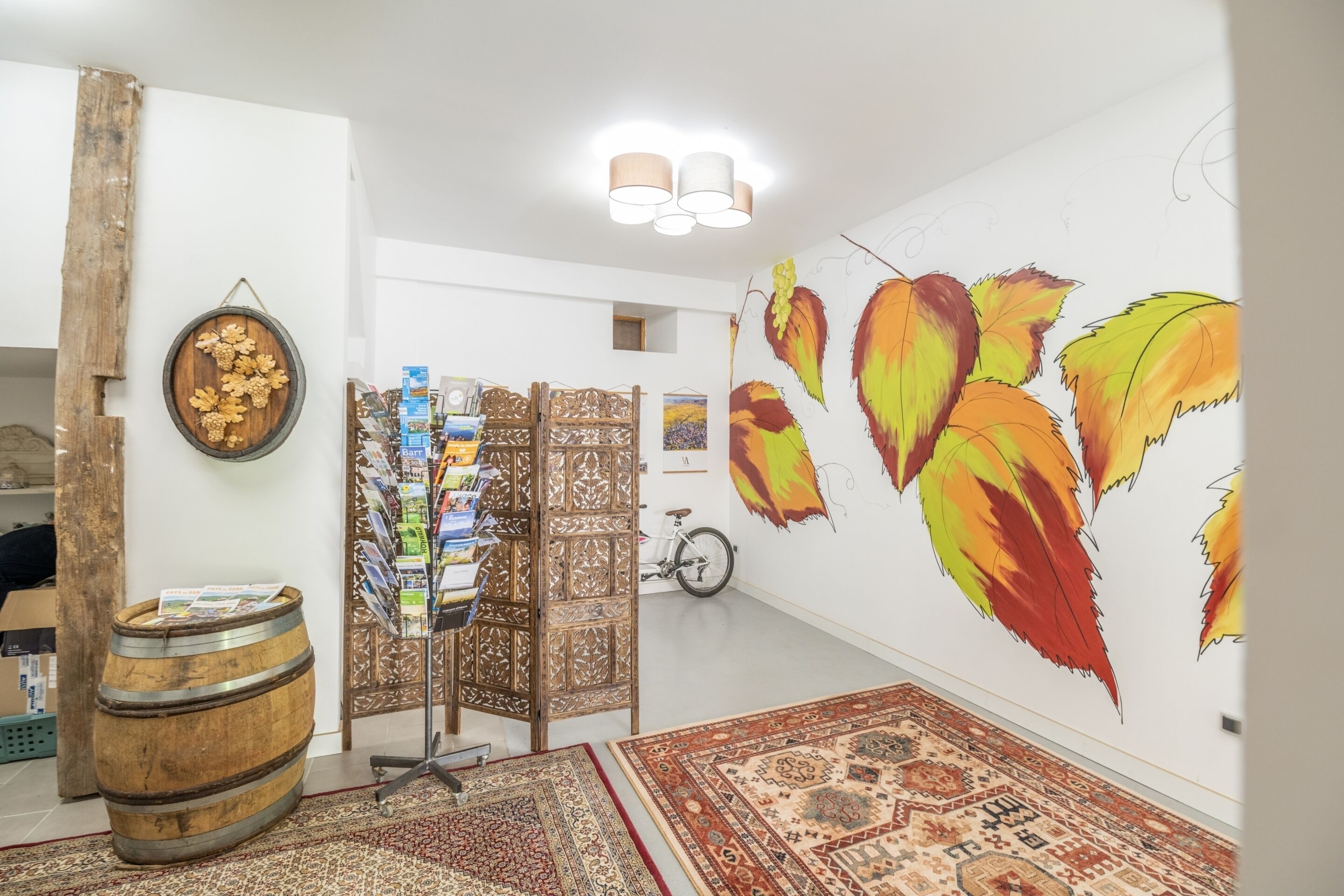 A room with a large wall mural, a wooden barrel, a patterned rug, and a decorative screen with magazines.
