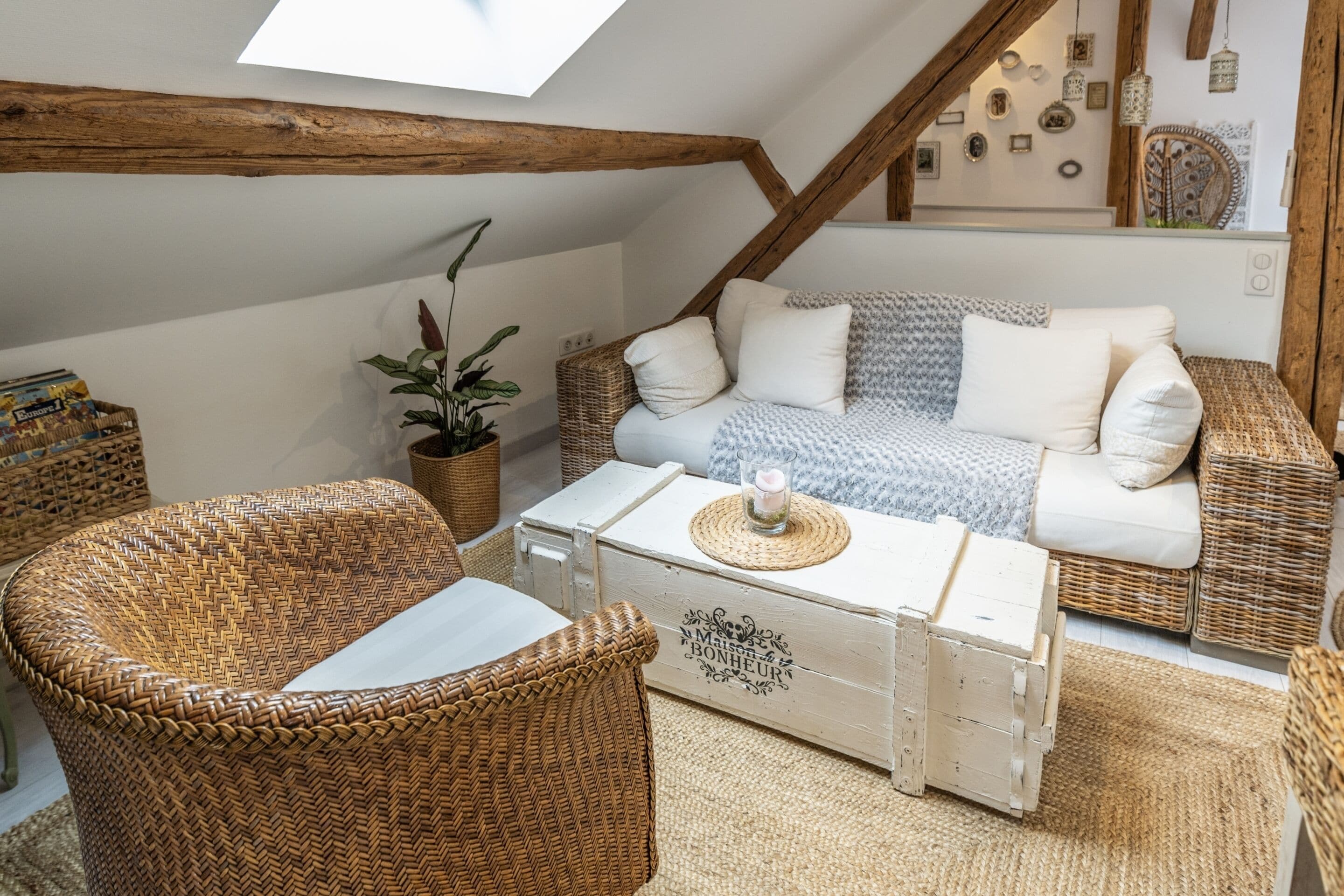 A cozy loft-style room with a high ceiling, exposed wooden beams, a white sofa with patterned cushions, a wicker armchair, and a small table with a candle.