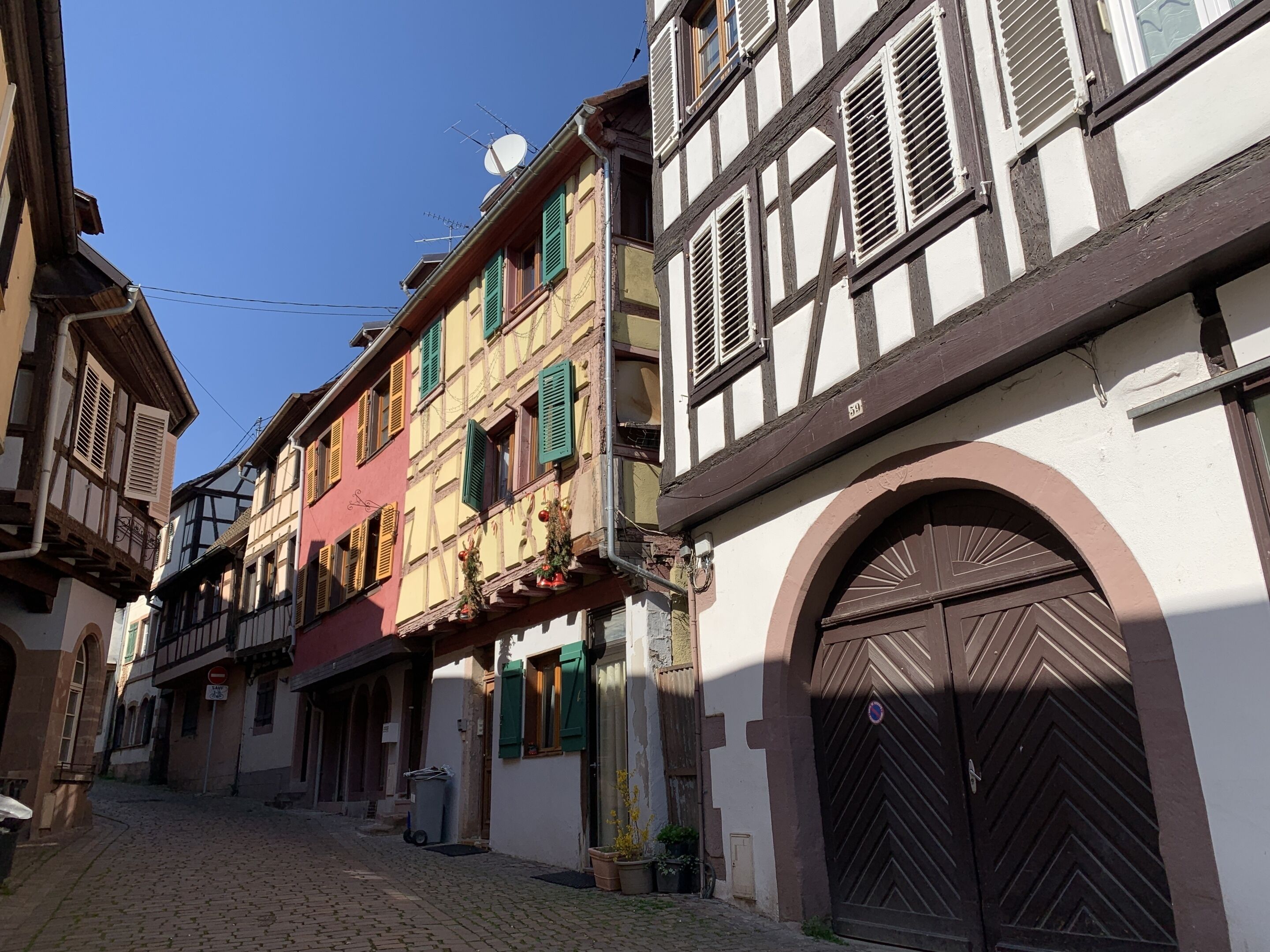 Une ruelle étroite pavée, bordée de bâtiments traditionnels à colombages, avec un mélange de volets ouverts et fermés, et une grande porte en bois en arc de cercle sur le côté droit.