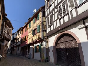 Una estrecha calle adoquinada flanqueada por edificios tradicionales con entramado de madera, con una mezcla de contraventanas abiertas y cerradas, y una gran puerta de madera arqueada a la derecha.