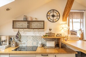 Une cuisine avec un plan de travail en bois, un étagère suspendue avec des bocaux, une horloge et une fenêtre avec des rideaux.