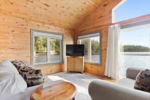 A cabin with wooden walls, a flat-screen TV on a wooden cabinet, and large windows overlooking a lake.