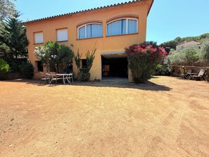 Una casa de dos pisos con cochera techada, zona de estar exterior y camino de tierra.