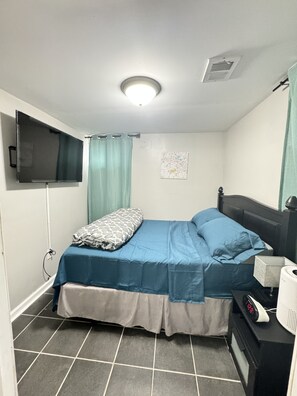 A small, neatly arranged bedroom with a bed, a flat-screen TV mounted on the wall, a bedside table with a lamp, and a clock. - Executive Apartment at Uplands (Baltimore)