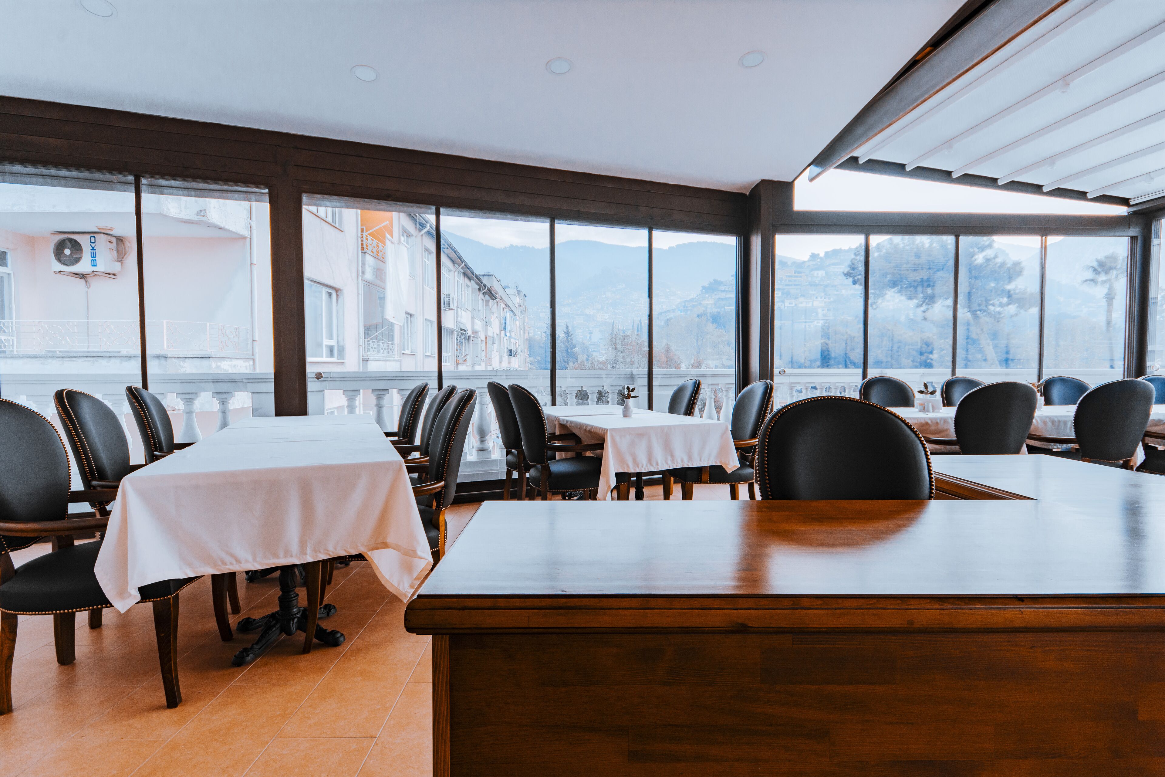 Um restaurante com janelas amplas, mesas com toalhas brancas e cadeiras pretas.