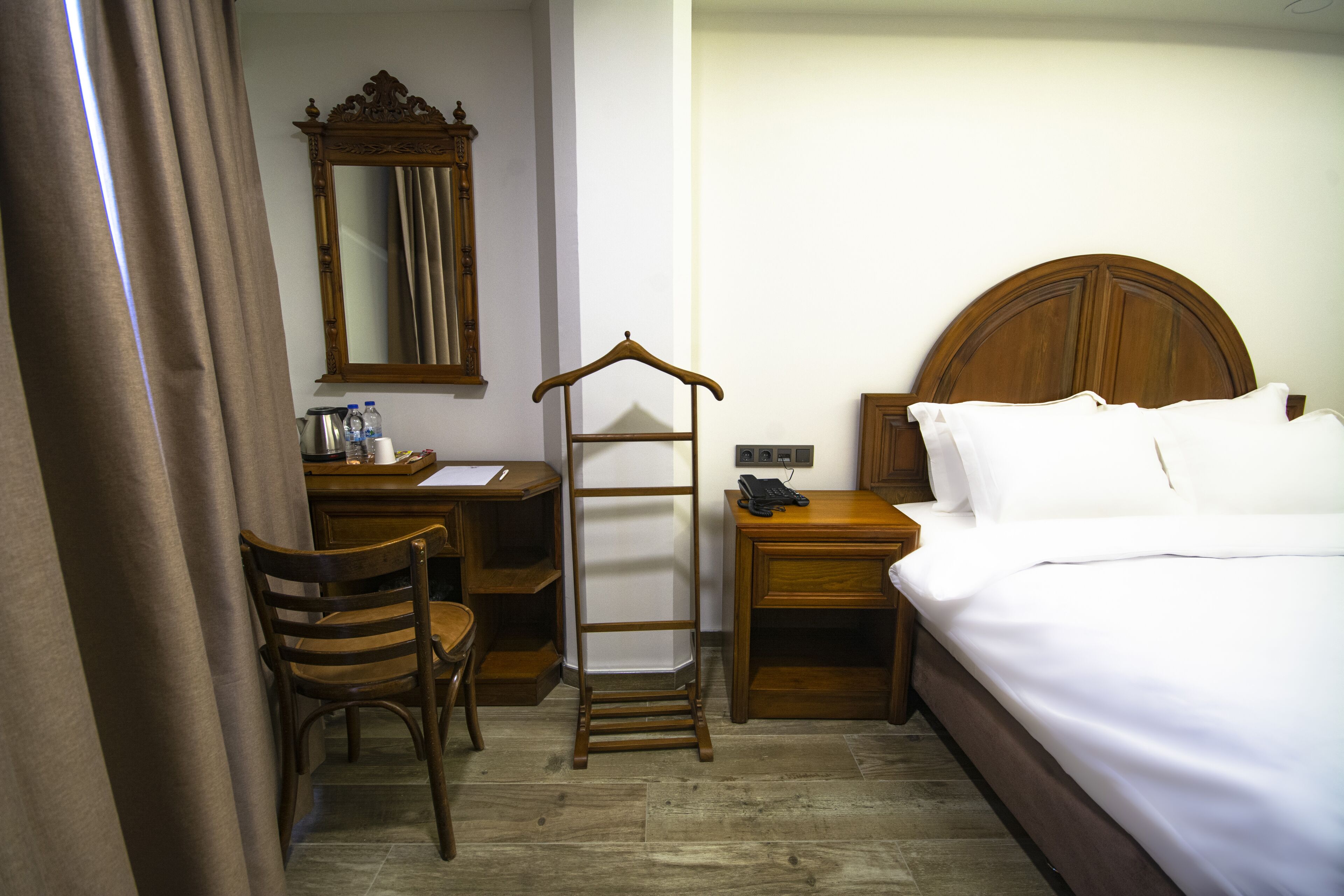 A hotel room with a wooden bed, a desk with a chair, a wardrobe, and a mirror on the wall.