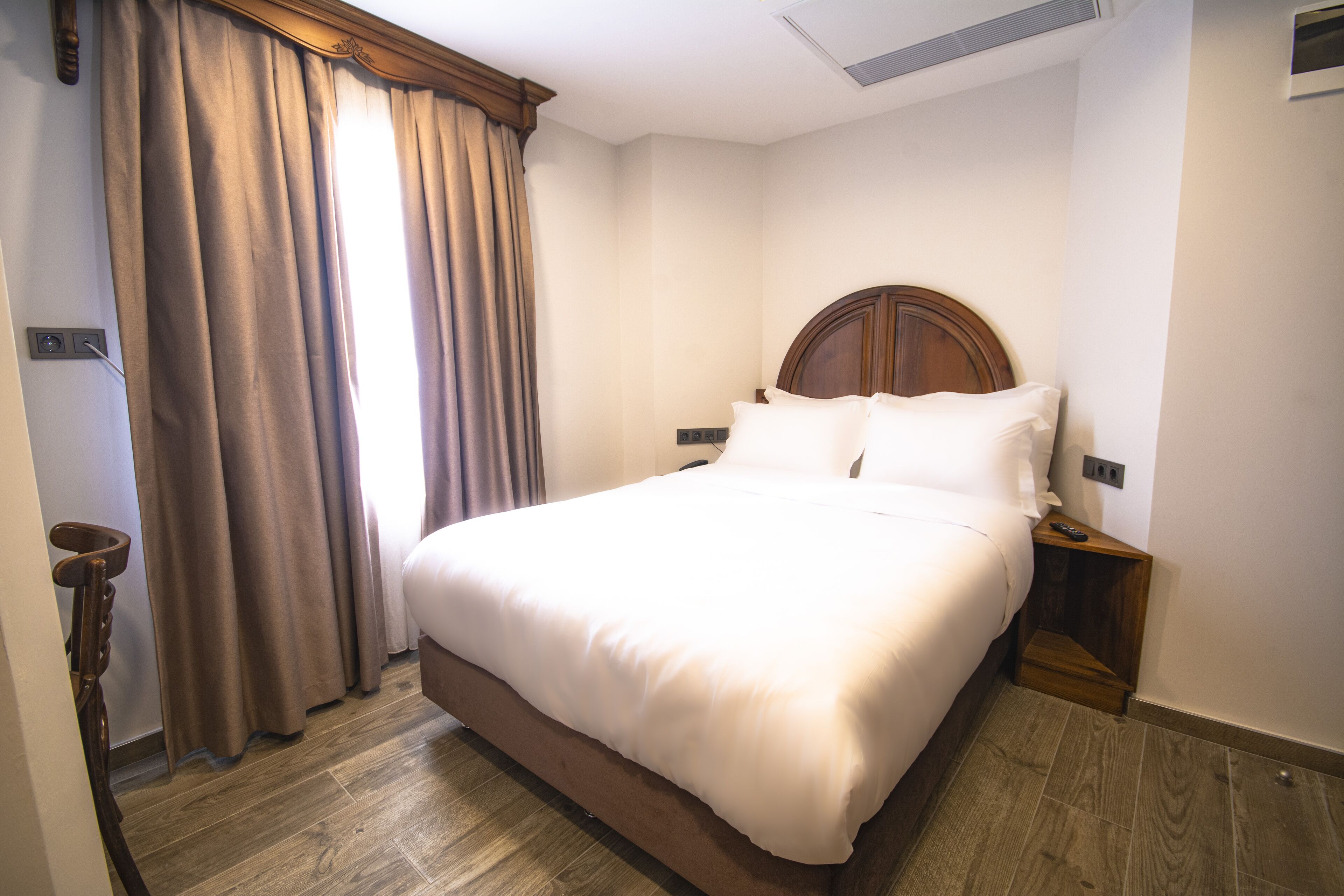 A neatly made bed with a wooden headboard, a bedside table with a lamp, and a window with curtains.