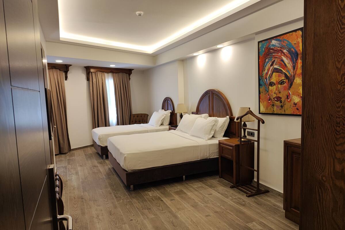 A hotel room with two beds, wooden flooring, a painting on the wall, and a ceiling light fixture.