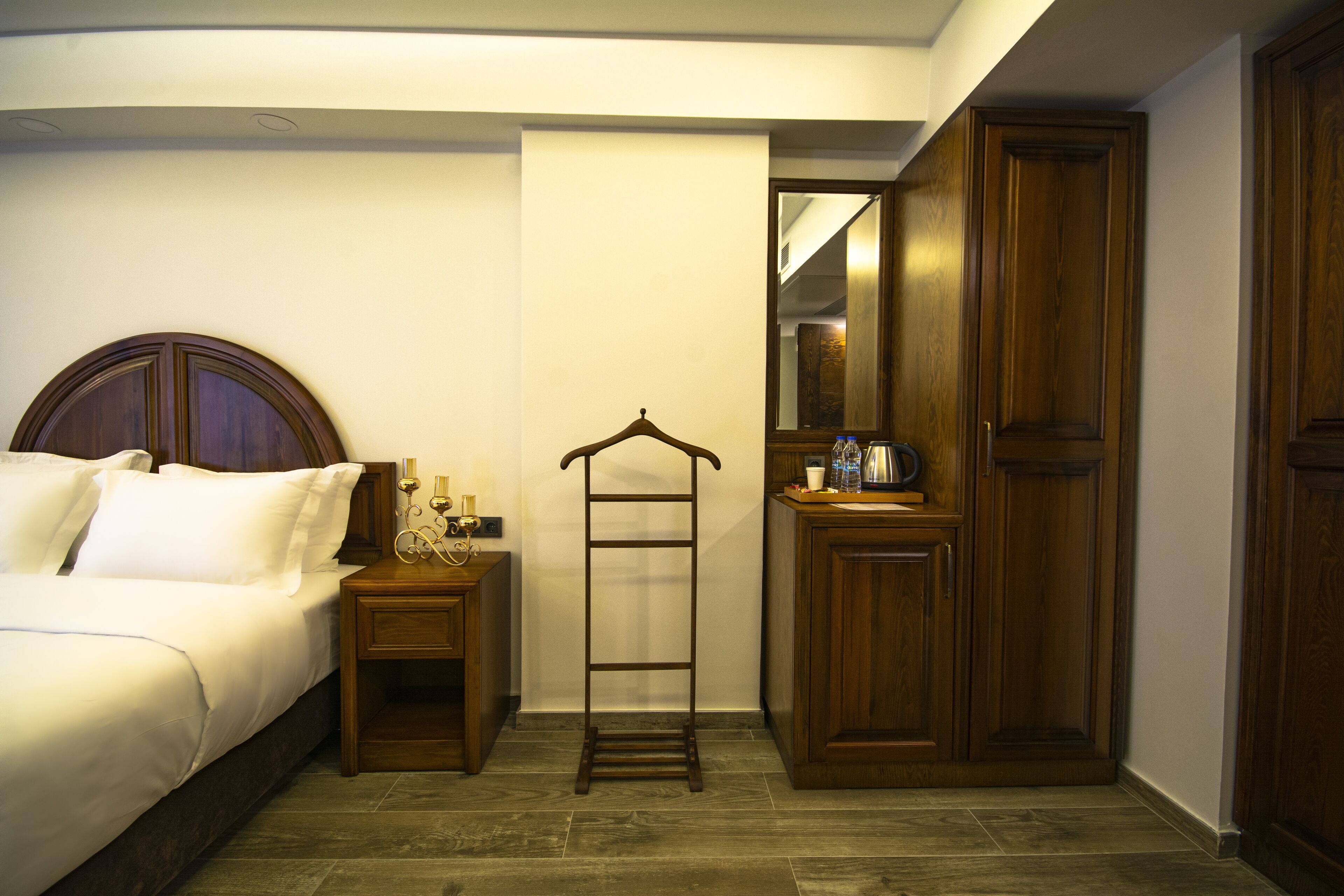 Een hotelkamer met een bed, nachtkastje, houten garderobe, spiegel en kledingroede.