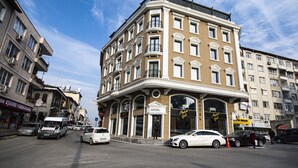 Um prédio de esquina com uma placa 'HOTEL', várias varandas e vista para a rua com carros e outros edifícios.