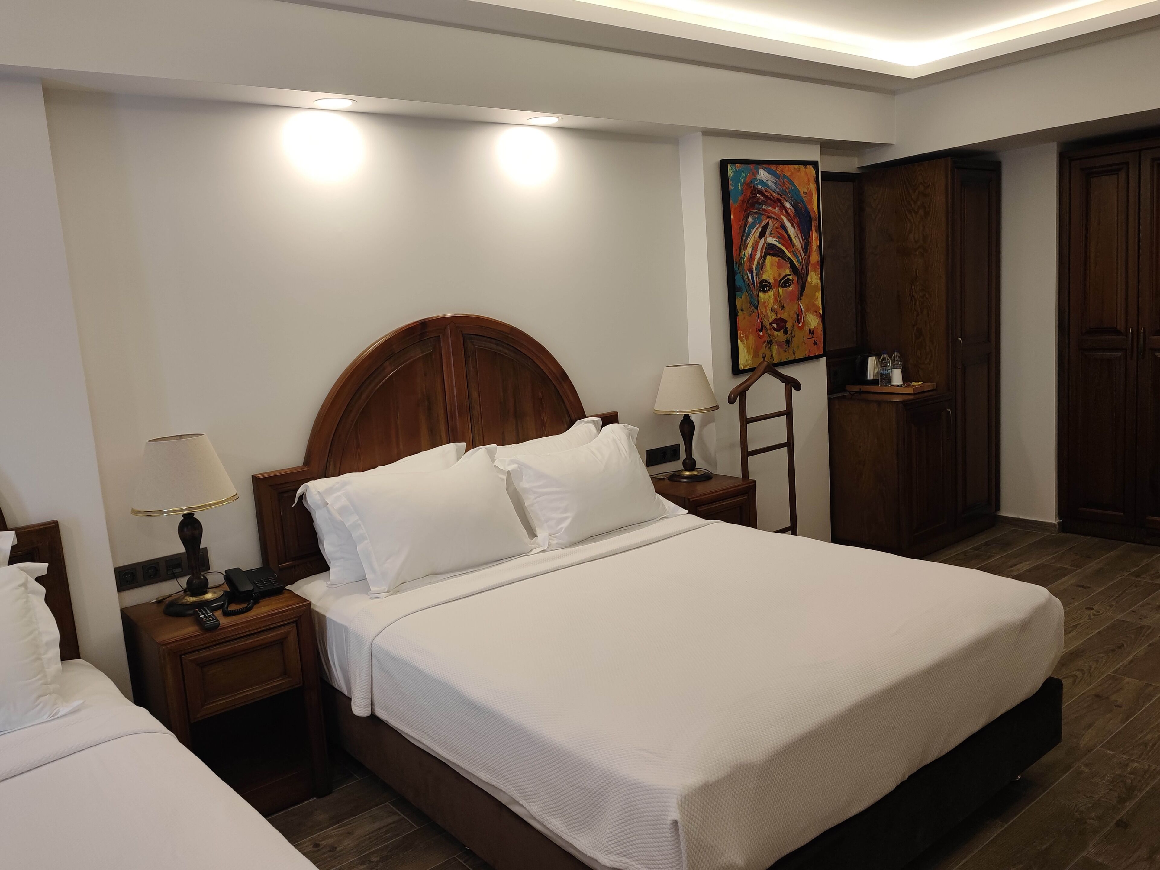 A hotel room with a large bed, two bedside tables with lamps, a wooden headboard, a painting on the wall, and a wardrobe.