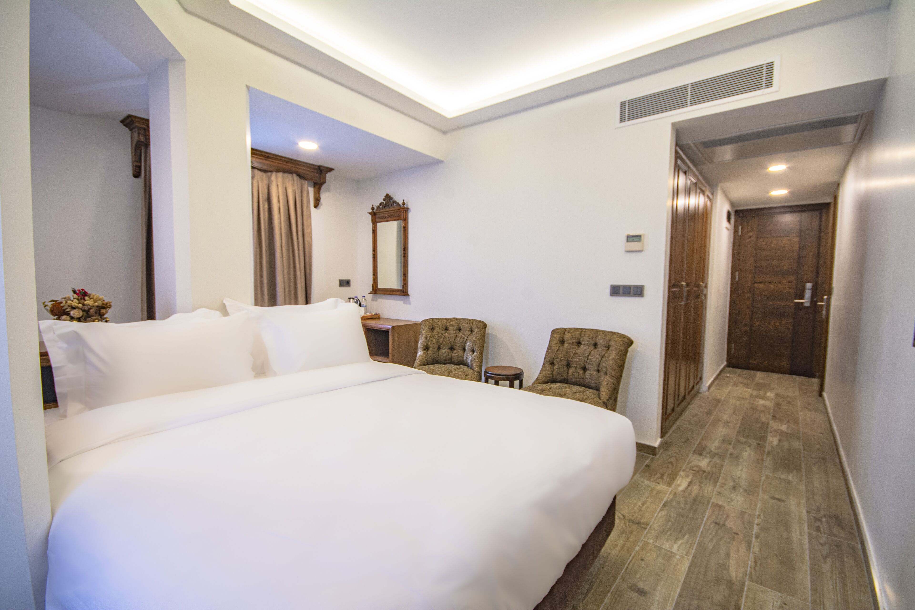 A hotel room with a large bed, two armchairs, a small table, a mirror, and a door leading to another room.