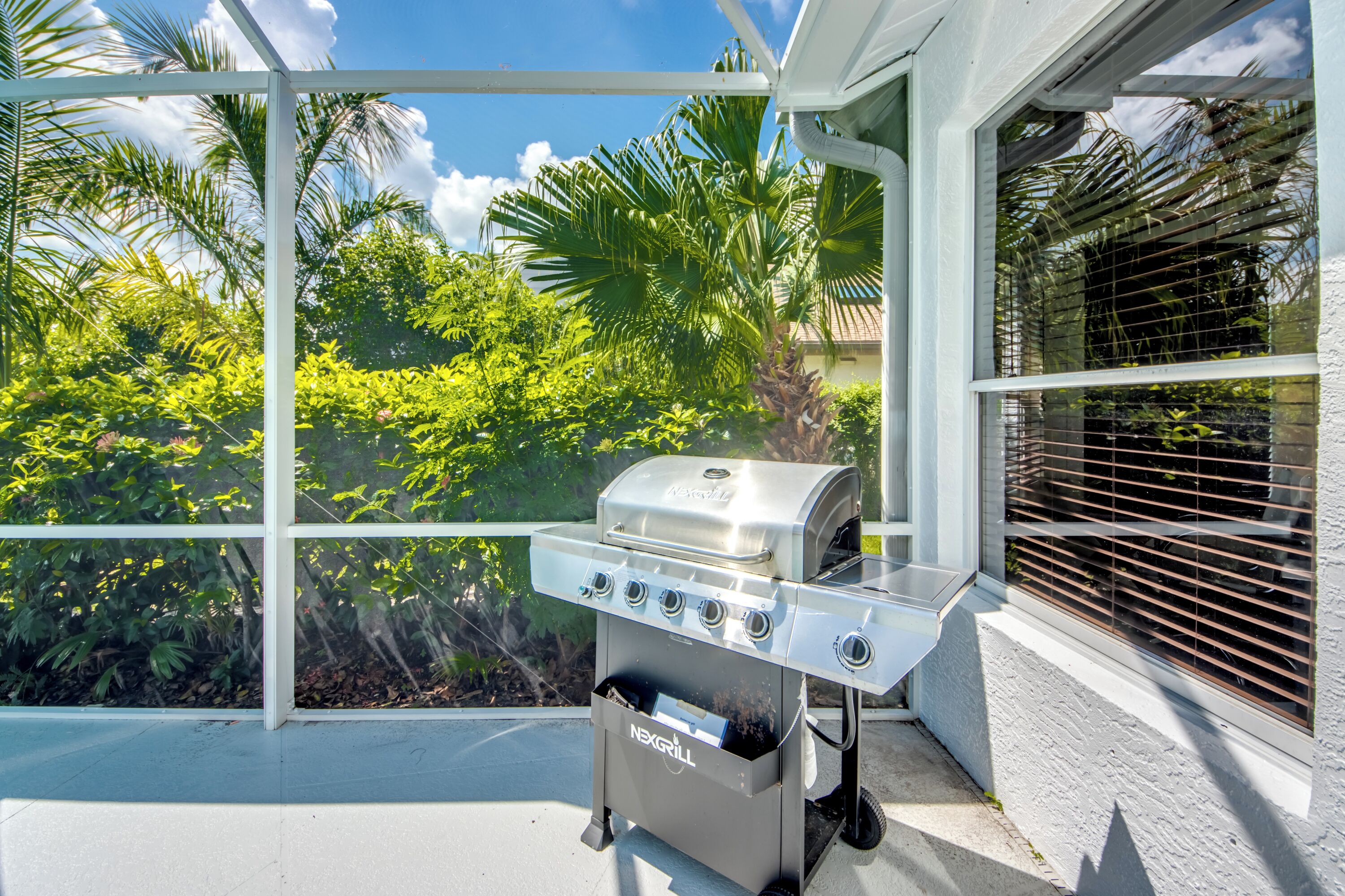 A patio with a stainless steel grill, a window with blinds, and lush greenery in the background.