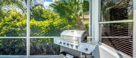 A patio with a stainless steel grill, a window with blinds, and lush greenery in the background.