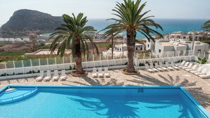 A pool area with lounge chairs and palm trees, overlooking a coastal landscape with buildings and a mountain. - Bluu Bahari EE (Karpathos)