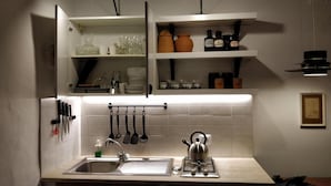 A kitchen with a sink, kettle, and hanging utensils. There are shelves above the sink with dishes and jars. - Casa Messi - Casa Messi a Vicolo Della Rivolta 31 Apt 2 (Pitigliano)